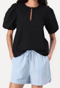 Kobieta ubrana w czarną bluzkę z bufkami i dekoltem z wycięciem oraz jasnoniebiesko-białe, paskowane spodenki z wiązanym sznurkiem, z rękami w kieszeniach.