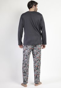 Hombre de pie descalzo, de espaldas, llevando una camiseta de manga larga oscura y pantalones a rayas en negro, blanco y rojo, sobre un fondo blanco.