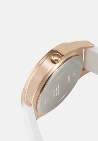 Roosa kuldne käekell külgvaates, kus on näha kroon, valge rihm ja roostevabast terasest tagakülg, millele on graveeritud brändi andmed.