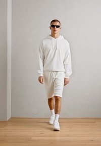 Hvit hettegenser og matchende shorts i et teksturert stoff. Brukt med hvite sneakers og mørke solbriller. Bakgrunnen er en lys grå vegg.