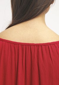 Röd, texturerad tyg med lös halsringning och draperade detaljer vid axlarna, som visar baksidan av en topp. Slät, subtil finish.