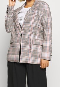 Kobieta ubrane w kolorową kurtkę w kratę na białym t-shircie z grafiką oraz czarnych szerokich spodniach, z ręką uniesioną do głowy.