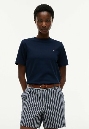 Donna che indossa una camicia blu navy a maniche corte infilata in pantaloncini a righe verticali blu navy e bianche con cintura marrone, in piedi con le mani dietro la schiena.