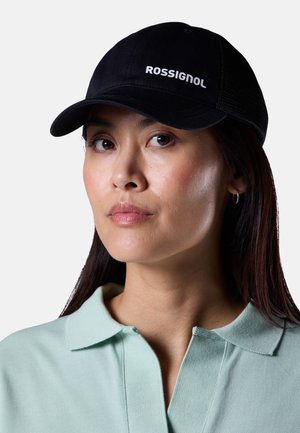 Donna con un cappellino nero Rossignol e una camicia verde chiaro con colletto, che guarda leggermente di lato su uno sfondo neutro.