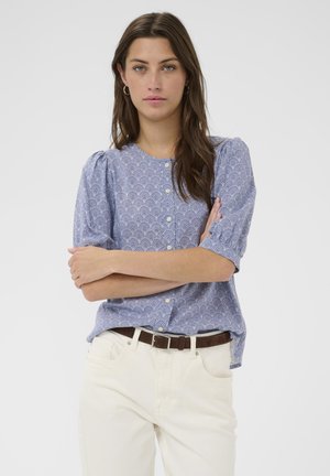 Kobieta o długich brązowych włosach, ubrana w niebieską, wzorzystą bluzkę na guziki, białe spodnie i brązowy pasek, stoi z założonymi ramionami.