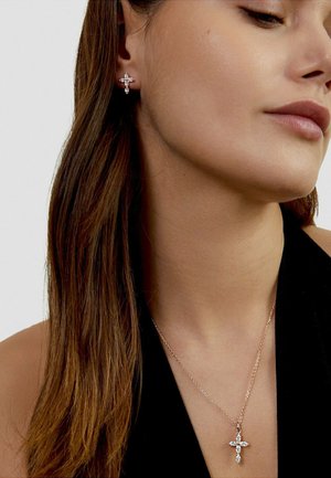 Vrouw met bijpassende kruisvormige diamantoorbellen en hangende ketting, met lang bruin haar en een zwarte mouwloze top.