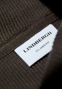 Dunkelbrauner Strickstoff mit geripptem Muster, ausgestattet mit einem weißen Etikett, auf dem "LINDBERGH" und "RELAXED FIT" in schwarzem Text zu lesen sind.