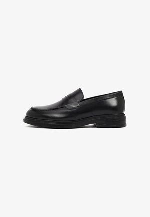 Mocasines de cuero negro con un acabado suave, punta redondeada y sutil detalle de costura. Presentan una suela de goma texturizada y baja, además de un diseño sin cordones.