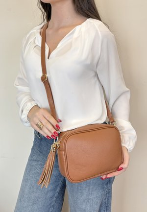 Donna con camicetta bianca e jeans blu tiene una borsa a tracolla in pelle color cuoio con dettaglio di nappine e ferrature dorate.