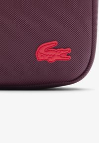 Borsa bordeaux con texture che presenta un logo in rilievo di coccodrillo rosso, con un bordo della cerniera liscio e angoli arrotondati.