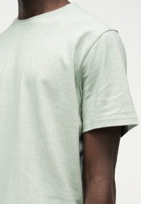 Κοντινό πλάνο ατόμου που φοράει απλό, ανοιχτό πράσινο κοντομάνικο μπλουζάκι με στρογγυλή λαιμόκοψη, μπροστά σε ουδέτερο φόντο.