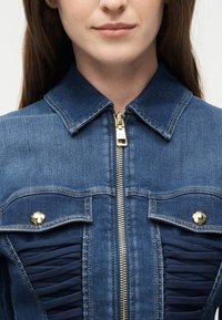 Γυναίκα που φοράει μπλε τζιν μπουφάν με χρυσό φερμουάρ και κουμπωτές τσέπες στο στήθος, εμφανιζόμενη από τους ώμους μέχρι τα χείλη.