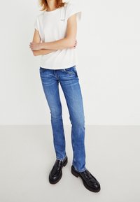 Camiseta blanca de manga corta con un pequeño logo, combinada con jeans azul de pierna recta y mocasines negros, sobre un fondo simple.