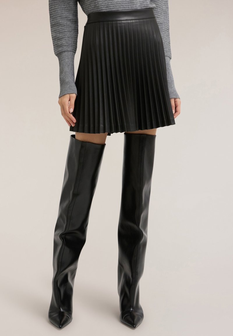 Svart plisserad kjol i läder med en figurnära midjeband, kombinerad med glänsande svarta stövlar som går över knäna, med spetsig tå.