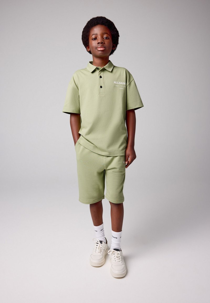 Lysegrøn polo med tre knapper og matchende shorts. Har en logo på trøjen og hvide sneakers med korte hvide strømper.