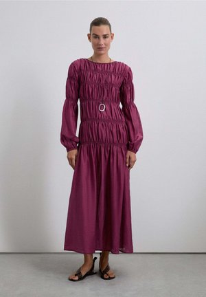 Vestido largo y fruncido en magenta profundo con mangas abullonadas, cintura elástica y un acento circular plateado. Llevado con sandalias planas negras.