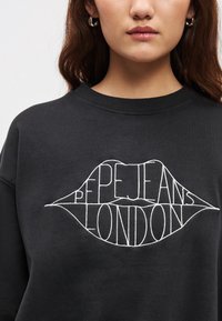 Μαύρο φούτερ με στρογγυλή λαιμόκοψη, διακοσμημένο με λευκή κεντητή επιγραφή σε σχήμα χειλιών: "PEPE JEANS LONDON." Απαλό βαμβακερό ύ�φασμα.