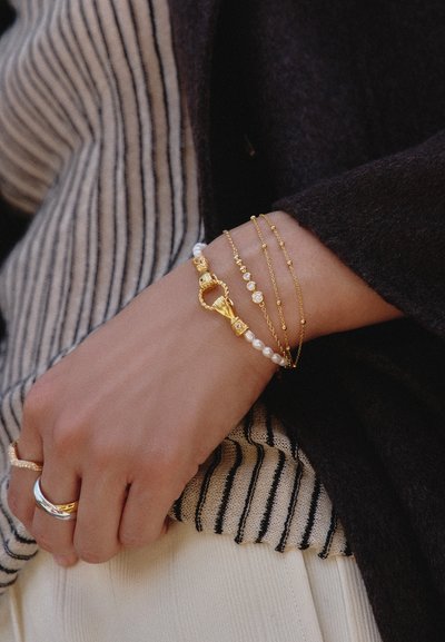 Πολύσειρα χρυσά και μαργαριταρένια βραχιόλια και χρυσά και ασημένια δαχτυλίδια στον καρπό, που ακουμπούν σε ριγέ και σκοτεινή υφασμάτινη ενδυμασία.