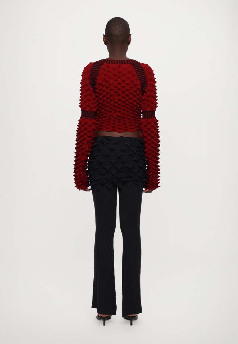 Roter strukturierter Pullover mit erhabenen Mustern, schwarzer Lagenrock und gerippten, dunklen Hosen, die einen Slim Fit und einen offenen Rückendetail aufweisen.