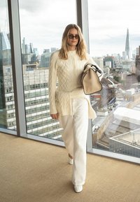 Naine valges soonikkoes kampsunis ja pükstes kõndimas toas, käes beež käekott, suured aknad tagaplaanil avavad linna silueti vaate.