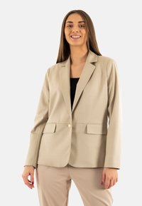 Blazer beige con solapas notch, dos bolsillos frontales, cierre de un solo botón y textura de tela suave. Combinado con pantalones beige.