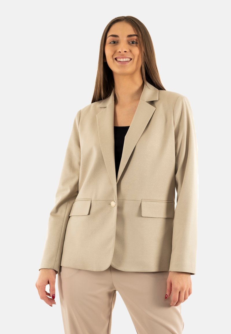 Blazer beige con solapas notch, dos bolsillos frontales, cierre de un solo botón y textura de tela suave. Combinado con pantalones beige.