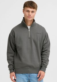 Sudadera gris con cuello alto, cierre de cremallera de cuarto y puños acanalados. Presenta el logo "A.R.C." en el pecho. Material suave.