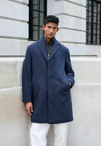 Marineblauer Wollmischungs-Mantel mit strukturierter Passform, breiten Revers und zwei Taschen, über einer schwarzen Zip-Jacke, kombiniert mit cremefarbenen Hosen.