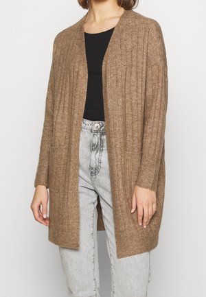 Personne portant un cardigan marron côtelé ouvert devant sur un haut noir et un jean taille haute gris clair sur fond blanc.