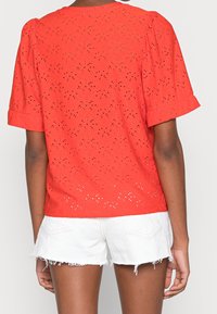 Top rouge à manches courtes avec motif en œillets, encolure ronde et manches légèrement bouffantes. Associé à un short blanc avec un ourlet effiloché.