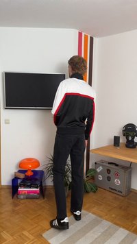 Športna črna trenirka z belimi in rdečimi poudarki, v kombinaciji s črnimi kavbojkami in črnimi čevlji z belimi črtami. Poleg je miza z oranžnim poudarkom.