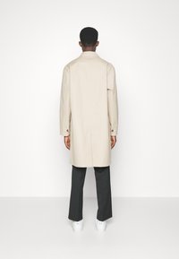 Mann, von hinten gesehen, trägt einen beigen langen Mantel, schwarze Hose und weiße Turnschuhe vor einem schlichten weißen Hintergrund.