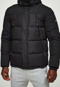 Černá prošívaná péřová bunda s kapucí, s kapsami na zip, prošívanou texturou a elastickými manžetami. Materiál je hladký a izolovaný.