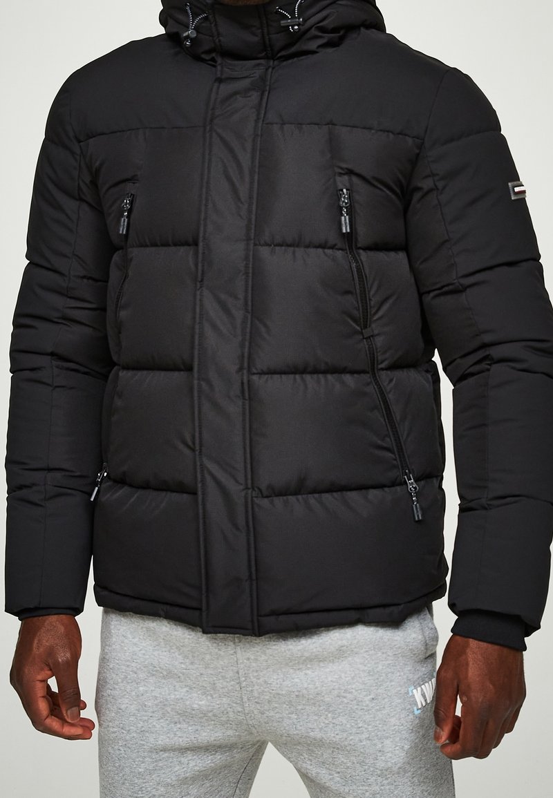 Černá prošívaná péřová bunda s kapucí, s kapsami na zip, prošívanou texturou a elastickými manžetami. Materiál je hladký a izolovaný.
