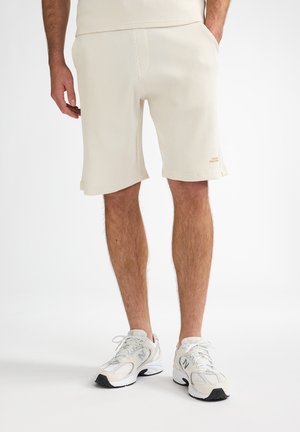 Crèmekleurige shorts met wafelhoedstructuur, zijzakken en een rechte snit. Gecombineerd met grijs-witte sportschoenen met zwarte accenten.