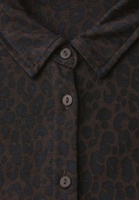 Camicia marrone scuro con colletto, decorata con motivo a leopardo nero e tre bottoni marroni visibili lungo la patta frontale.