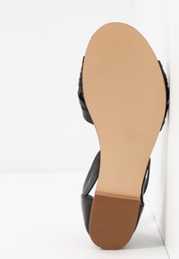 Mustat sandaalit, joissa on punottuja hihnoja, litteä pohja ja teksturoitu päällinen. Pohja on beige ja siinä on uurretta pitoa varten. Minimalistinen muotoilu, ilman koristeita.