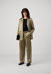 Licht olijfgroene op maat gemaakte blazer en bijpassende cargobroek met een gladde textuur, gecombineerd met een zwarte top en leren zwarte schoenen.