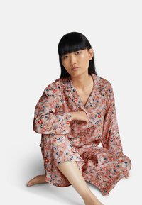 Giovane donna con capelli neri che indossa pigiama floreale rosa, seduta con le gambe incrociate su un pavimento bianco contro uno sfondo bianco semplice.