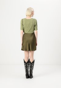Top verde plissado de mangas curtas combinado com uma saia mini de cetim verde-oliva e botas pretas de estilo western com detalhes intrincados em prata.