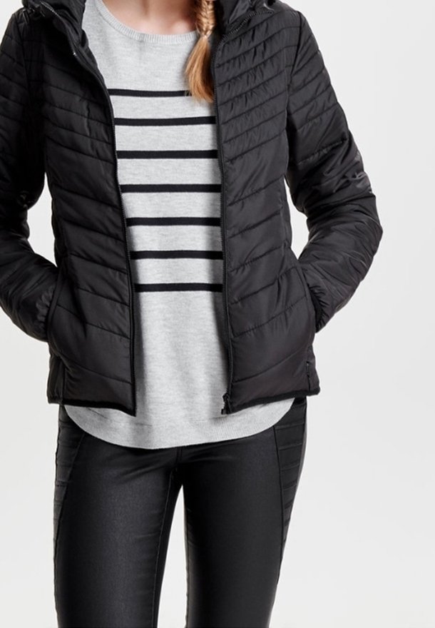 Personne portant une veste matelassée noire sur un pull gris avec des rayures horizontales noires et un pantalon noir, les mains dans les poches de la veste.