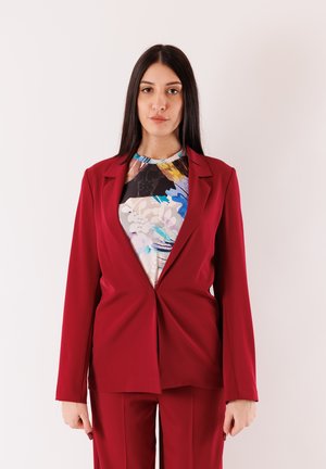 Blazer rosso su misura con chiusura singola, abbinato a una maglia con motivo multicolore. Tessuto liscio, spalle strutturate e maniche lunghe.