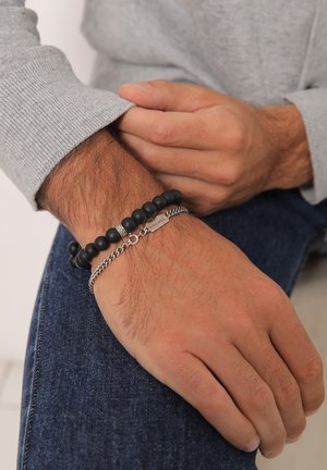 Black bead bracelet with silver accents, paired with a silver chain bracelet featuring a small tag. Worn on a wrist with light skin tone.
