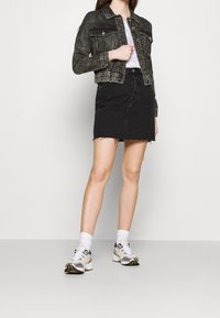 Žena v outdated černé džínové bundě, bílém tričku, černé džínové sukni, bílých ponožkách a bílých sportovních teniskách na jednotném pozadí.