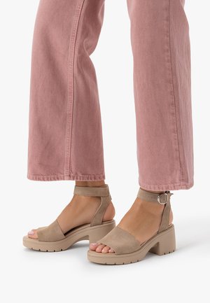 Stopy w beżowych sandałach na masywnym obcasie z paskami wokół kostki, zestawione z krótkimi, szerokimi spodniami w kolorze przybrudzonym różowym.