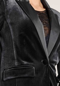 Zwart fluwelen blazer met een satijnen revers, enkele knoopsluiting en een voorkant zak. Gladde textuur met subtiele glans.