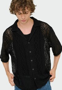 Camisa negra de encaje con botones y mangas cortas, que presenta un patrón geométrico de tejido abierto y un cuello, llevada sobre una camiseta sin mangas negra.