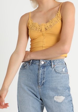 Vrouw in een mosterdgele, met kant afgewerkte, mouwloze cropped top en lichtblauwe, hooggetailleerde gescheurde jeans, staand met één arm gekruist.