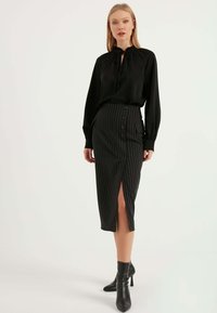 Svart långärmad blus med knytband; åtsittande svart pennkjol med vita randningar, sidöppning och knappelement. Svarta klackkängor.