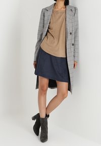 Manteau long gris à carreaux avec un design croisé à double boutonnage, assorti à un haut beige et une jupe en daim marine. Des bottines grises complètent la tenue.